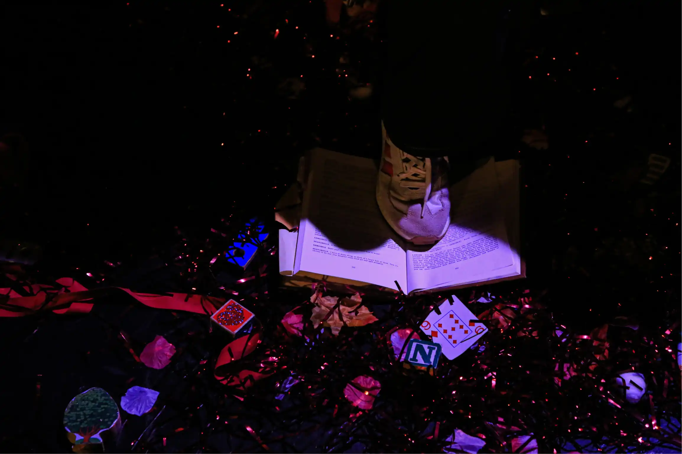 A shoe steps on a printed copy of 'The Cherry Orchard,' which sits on a black floor covered with confetti, playing cards and wood blocks