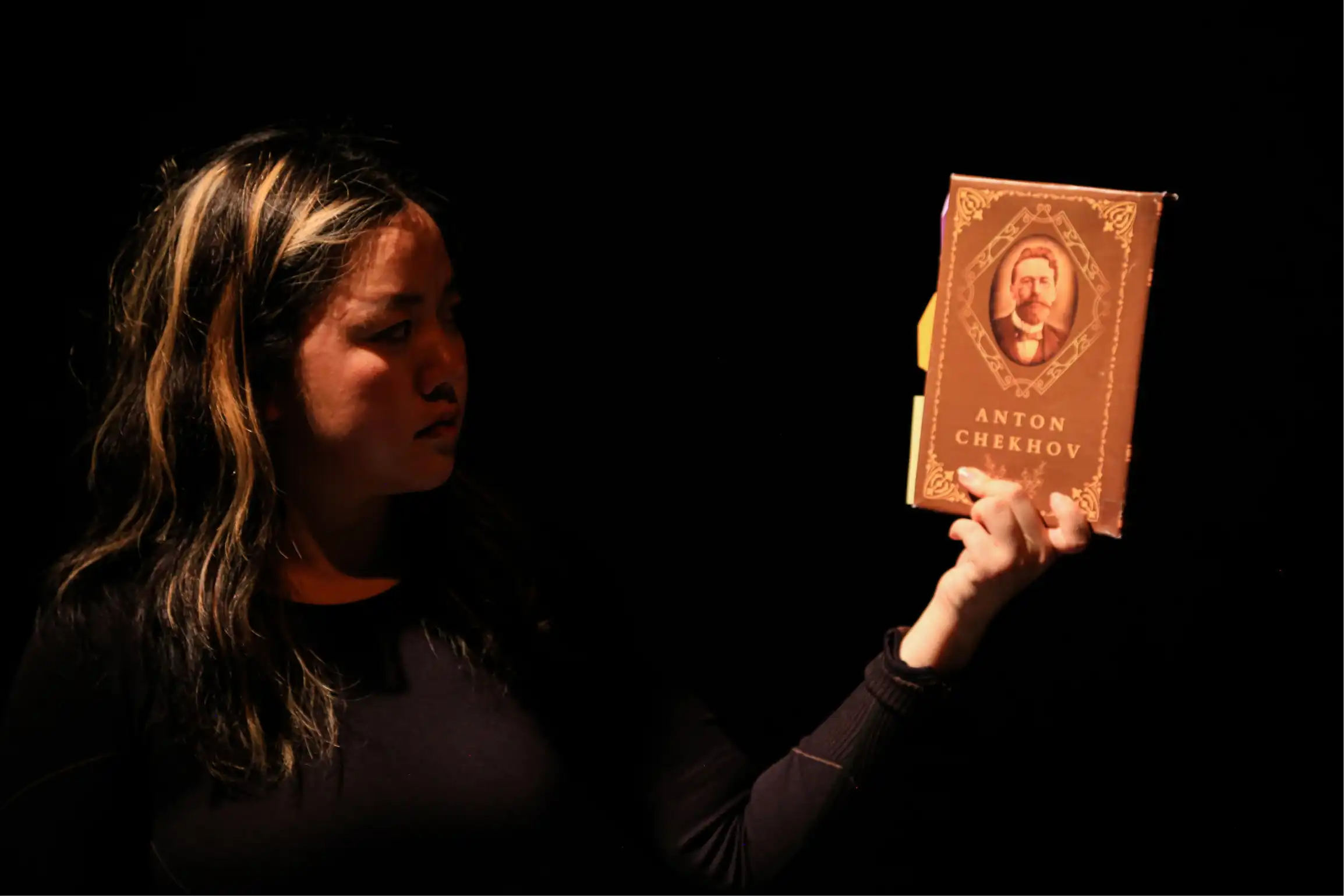 Ariel holds a printed copy of Anton Chekhov's 'The Cherry Orchard' under bright lights against a black background